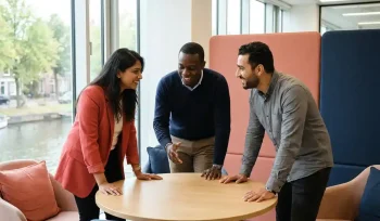 Drie diverse professionals in gesprek aan een ronde tafel in een modern, licht Nederlands kantoor met koraalrode en marineblauw accenten.