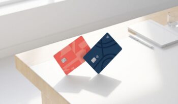 Two payment cards with coral red and navy blue accents on a clean wooden desk in bright natural light.