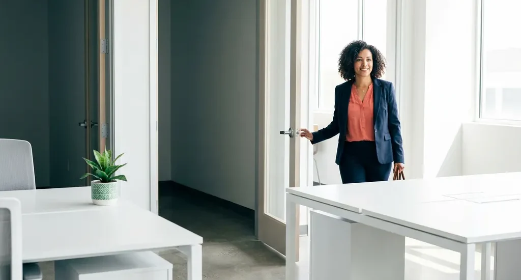 Zelfverzekerde vrouw die een lichte, moderne werkruimte betreedt via een open deur, met een groene plant op een wit bureau.