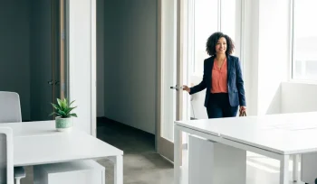 Zelfverzekerde vrouw die een lichte, moderne werkruimte betreedt via een open deur, met een groene plant op een wit bureau.